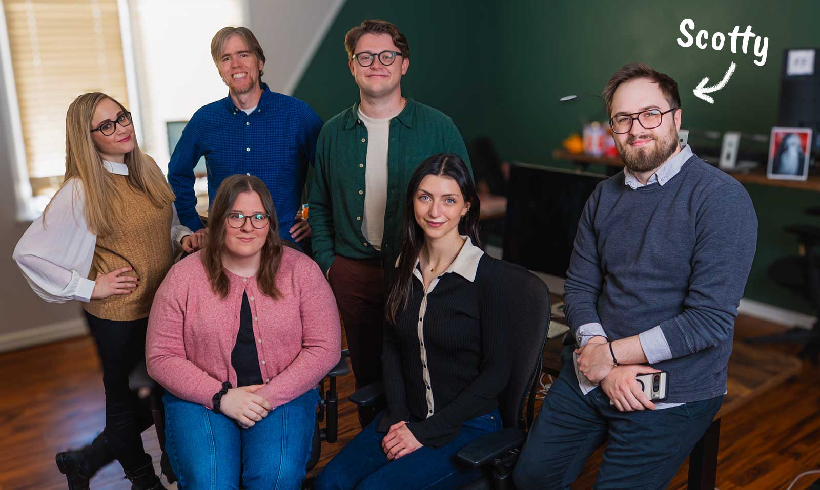 Six people pose and smile in an office setting. One man on the right, labeled “Scotty” with an arrow, holds a phone. The group sits or stands in front of desks, with a window and wooden floor in the background.
