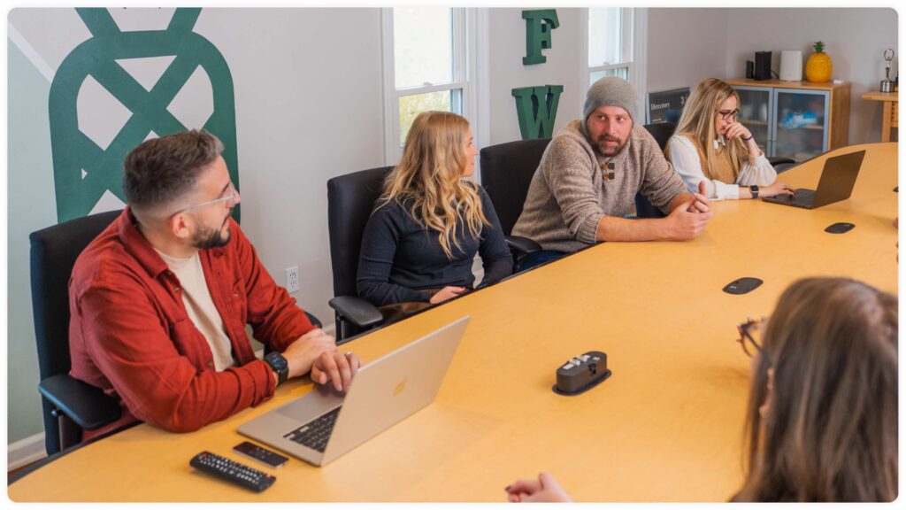 Five people sit around a conference table in a modern office, engaged in discussion. Laptops, a remote, and other devices are on the table. A green abstract wall design and large windows are visible in the background.